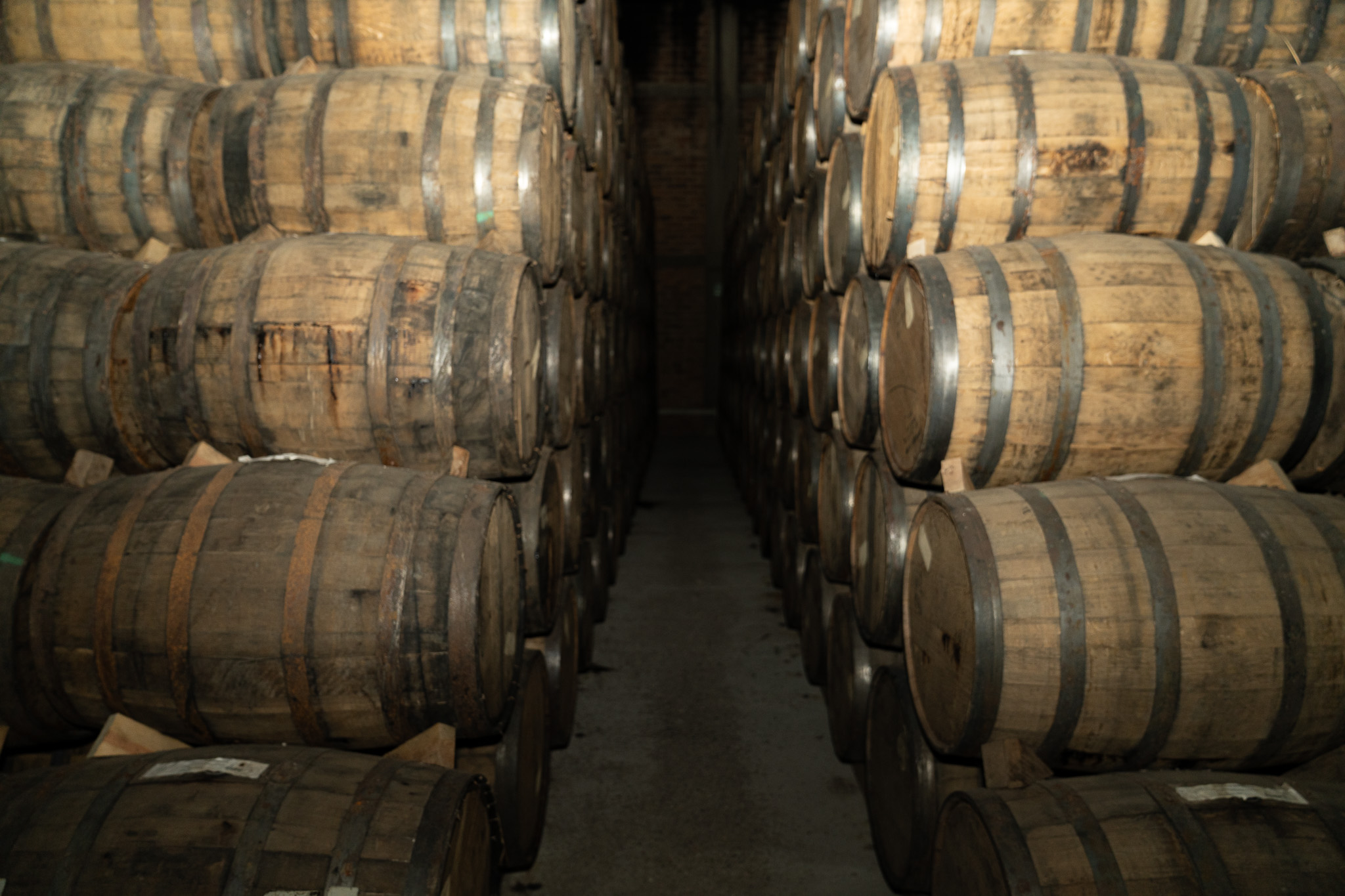 A corridor between stacked oak barrels in a dim rickhouse, warm light on the staves.