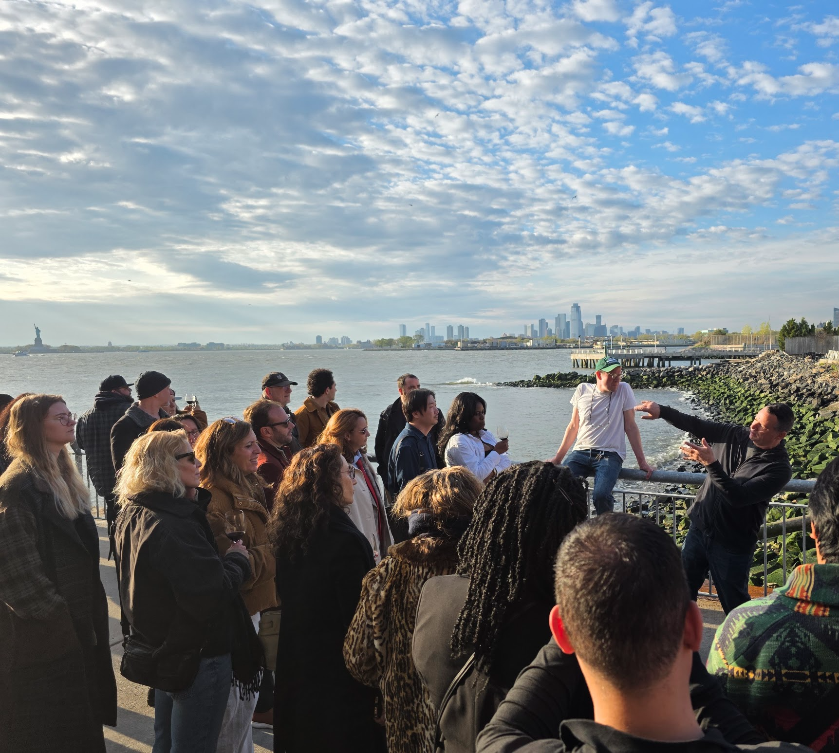 A crowd gathered along a waterfront railing at golden hour, harbour and city skyline behind them, one host mid-gesture addressing the group.